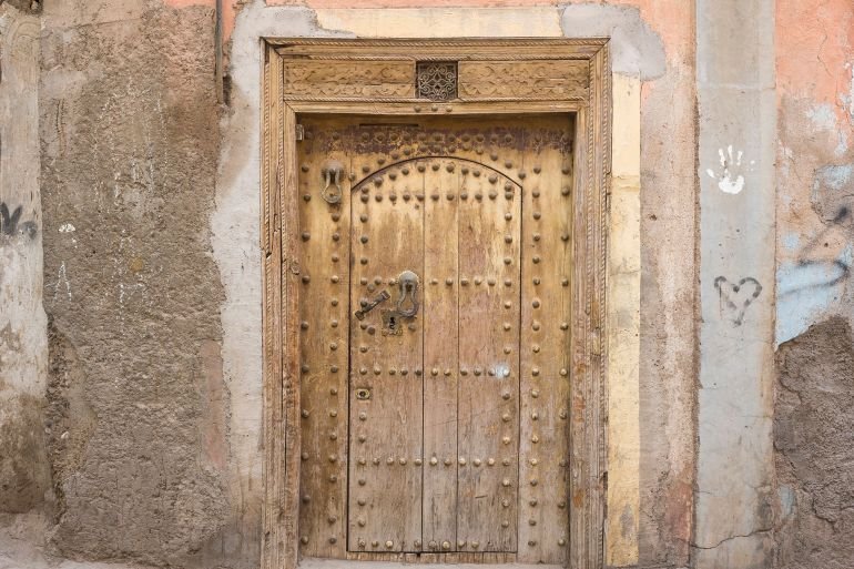 Marrakech, Morocco – May 3, 2025: Antique wooden door with carved architrave in medina. Weathered pink wall, rivets, ornate keyhole, traditional door knocker. Authentic Moroccan architectural detail; Shutterstock ID 2624172317; purchase_order: aj; job: ; client: ; other: