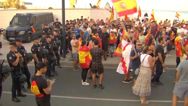 Law enforcement personnel watch while protesters hold Spanish flags, as Ceuta residents destroy migrant camp by the beach, in Ceuta, Spain, August 27, 2026, in this screen grab taken from a handout video. FORTA/Handout via REUTERS THIS IMAGE HAS BEEN SUPPLIED BY A THIRD PARTY. SPAIN OUT. NO COMMERCIAL OR EDITORIAL SALES IN SPAIN