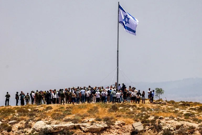 Israeli settlers, accompanied by Israel's far-right finance minister Bezalel Smotrich, raise a large Israeli flag during a cornerstone laying ceremony for a new settlement on Mount Tarousa, near the town of Dura west of Hebron in the occupied West Bank, on June 16, 2026. (Photo by HAZEM BADER / AFP via Getty Images)