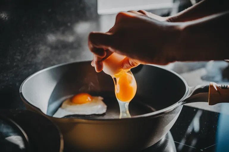 Close-up of female hand cracking an egg into a sizzling hot frying pan as she prepares a healthy breakfast in kitchen in the morning. Clean eating and healthy eating habits with natural ingredients. Wellness lifestyle and mindful meal preparation