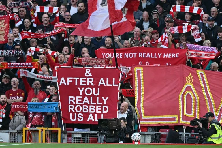 Liverpool fans hold signs mocking Manchester City's financial scandal as the television crew work pitch-side ahead of the English Premier League football match between Liverpool and Manchester City at Anfield in Liverpool, north west England on October 11, 2026.